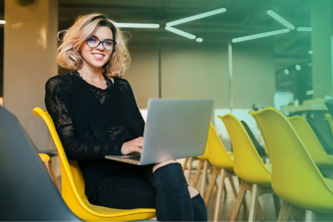 Mujer rubia sentada en una silla amarilla y usando su portátil en una habitación grande