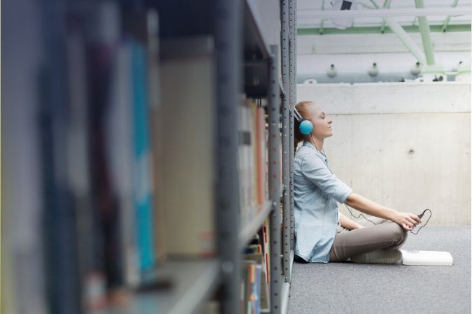 Mujer escuchando música sentada junto a una estantería en una biblioteca