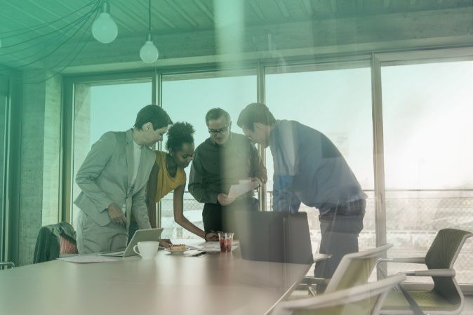 4 personas participan en una reunión de grupo en una oficina con grandes ventanales